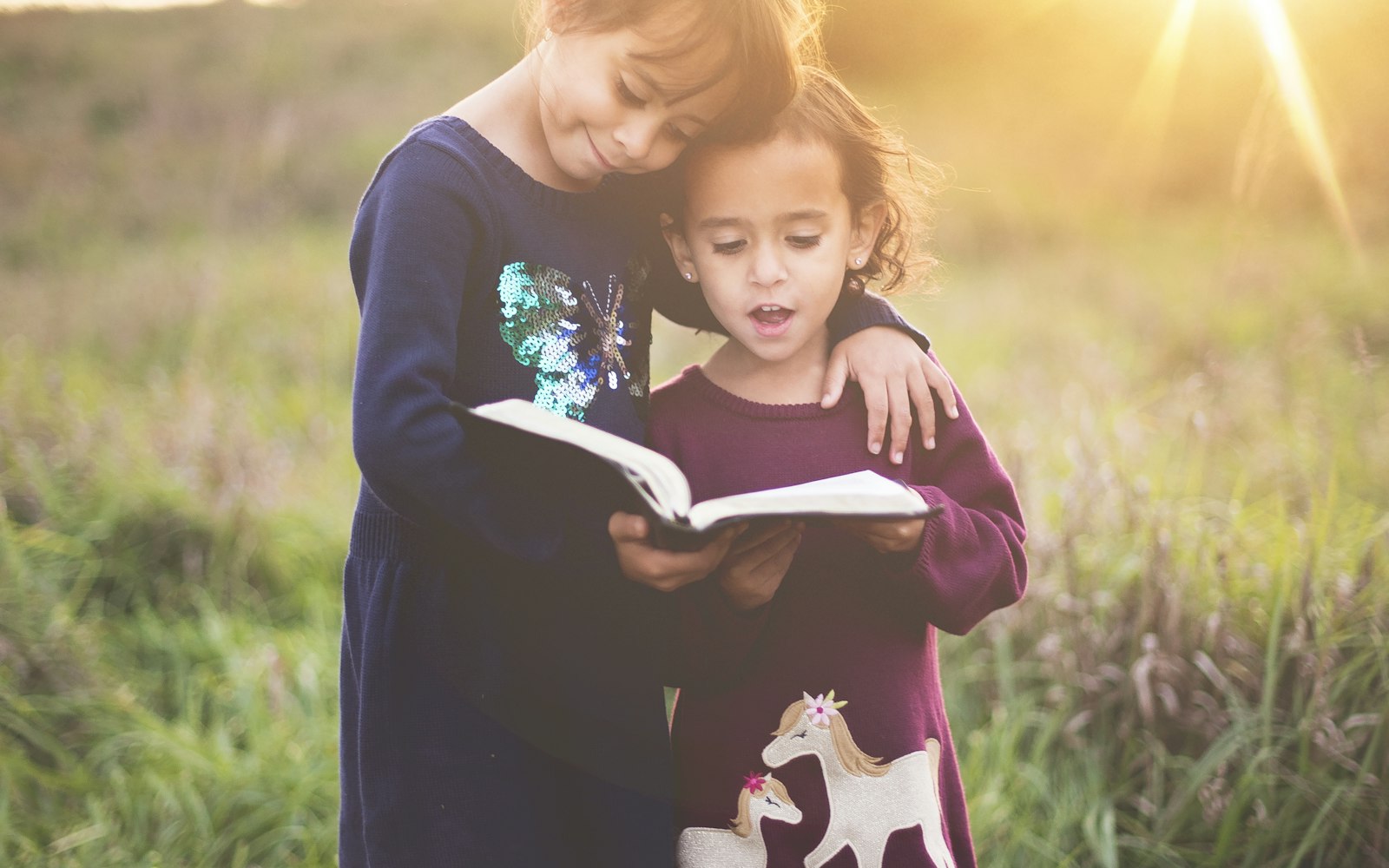 A child reading a book with an older sibling, sunlight behind them